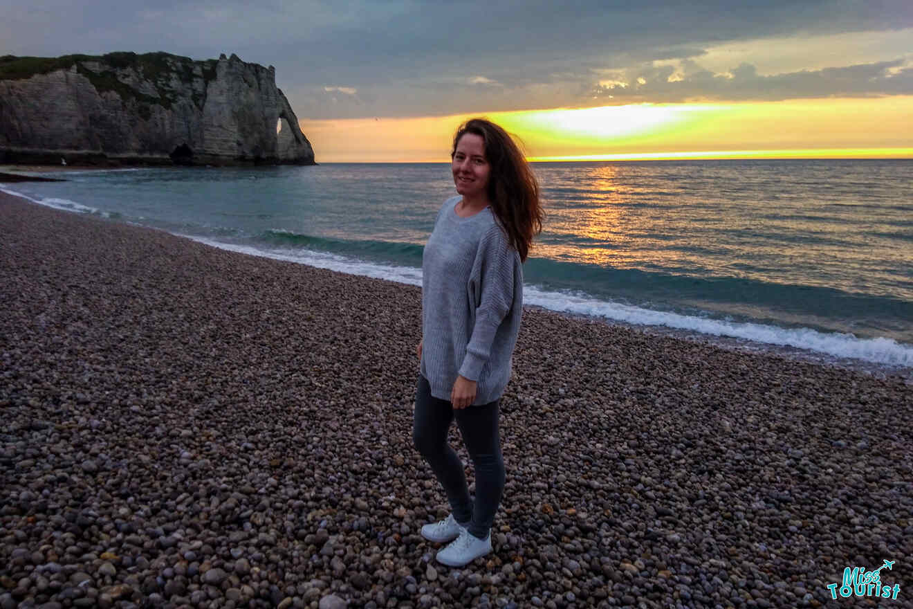 The writer of the post standing on a pebble beach with cliffs in the background and a sunset over the ocean.