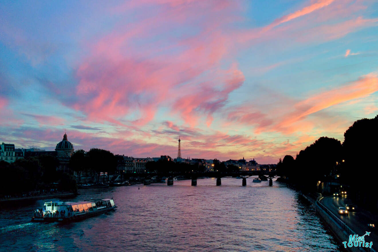 Sunset over a river with a boat, silhouetted bridge, and city skyline in the background, beneath a sky filled with colorful clouds.