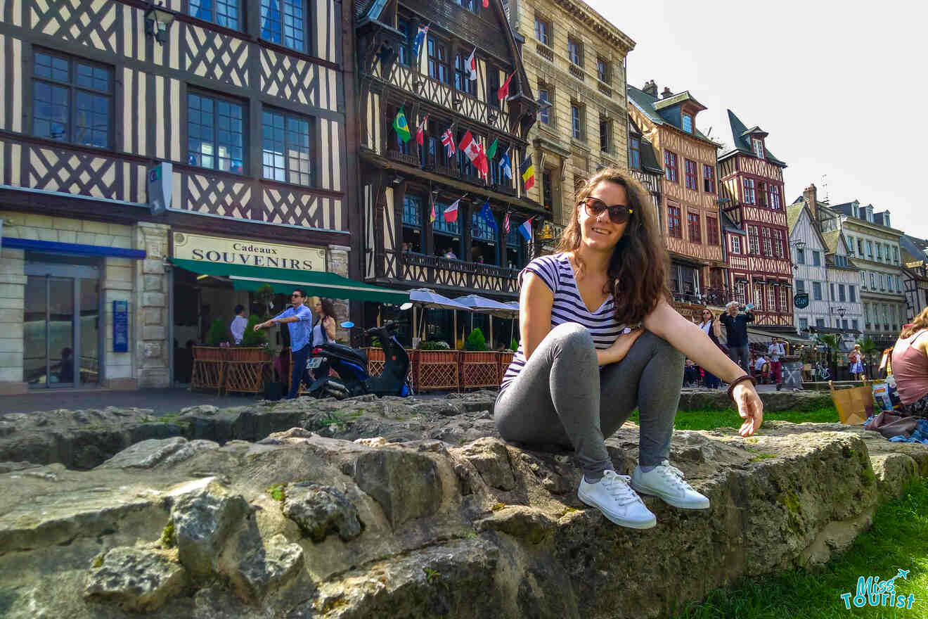 The writer of the post sitting on a stone wall in front of colorful half-timbered buildings with a souvenir shop and outdoor seating area in a lively street scene.