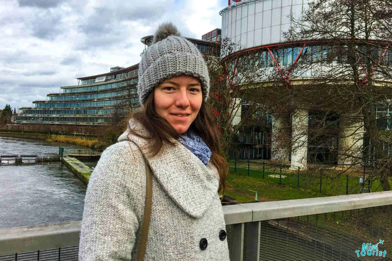 The writer of the post in a gray coat and knit hat stands on a bridge near a modern building and river.