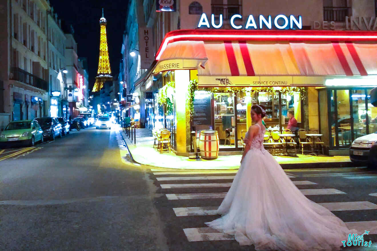 A bride in a wedding dress stands on a Parisian street at night, near a brightly lit café. The Eiffel Tower is visible in the background.