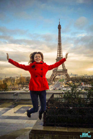 The writer of the post in a red coat jumps joyfully in front of the Eiffel Tower on a cloudy day.