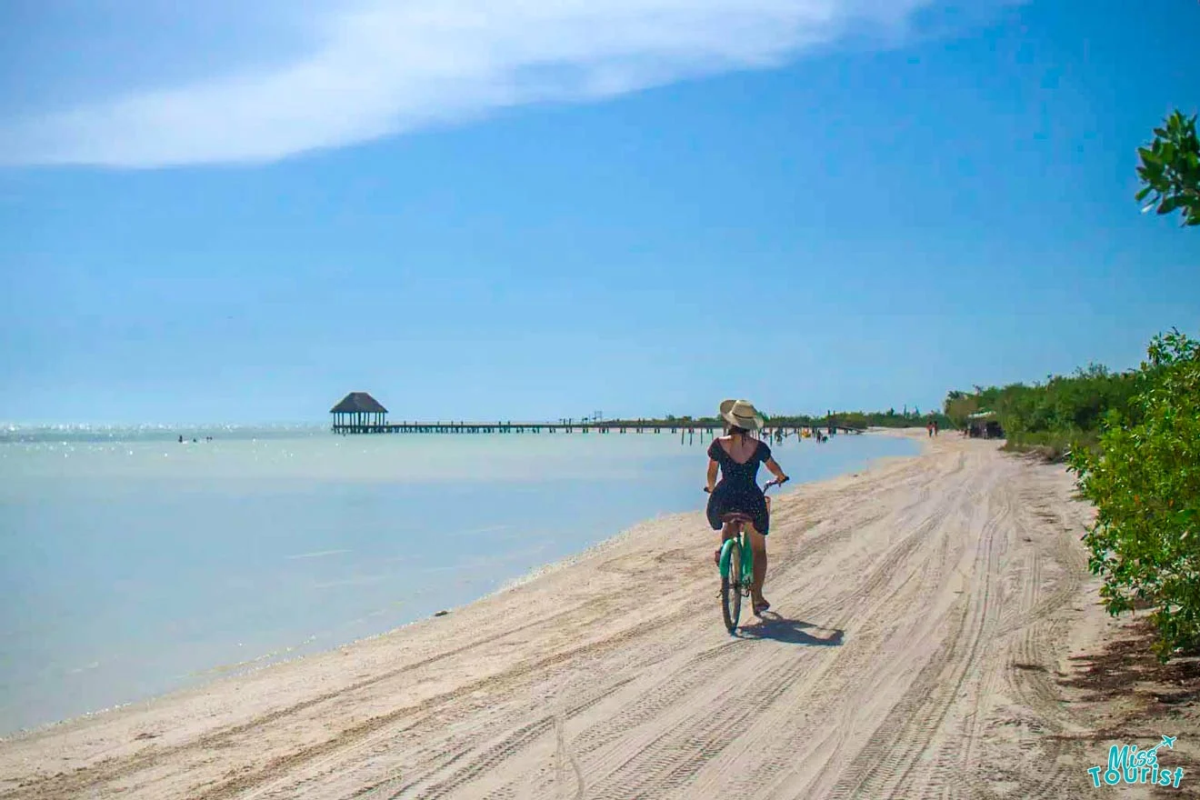 The writer of the post rides a bicycle on a sandy beach with clear blue skies. A pier extends into the water in the background.