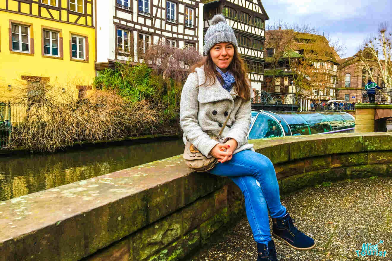 The writer of the post wearing a gray coat and hat sits on a stone wall by a canal, with traditional half-timbered houses in the background.