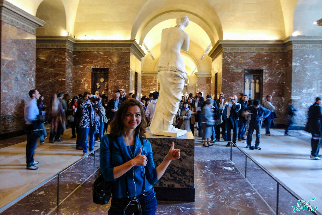 The writer of the post poses smiling with thumbs up in front of the Venus de Milo statue at a museum, surrounded by a crowd of people.