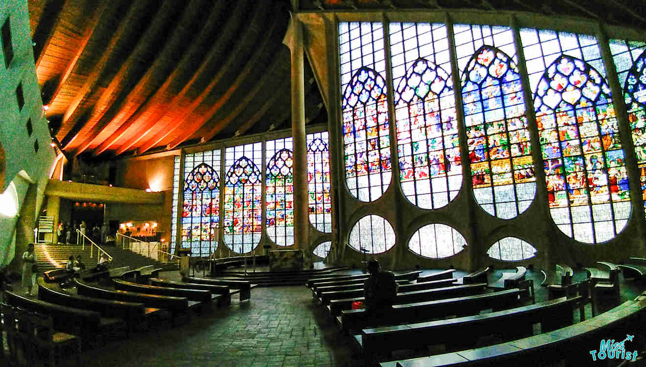 Interior of a church with large, vibrant stained glass windows on the curved walls and wooden pews arranged in a semicircle.