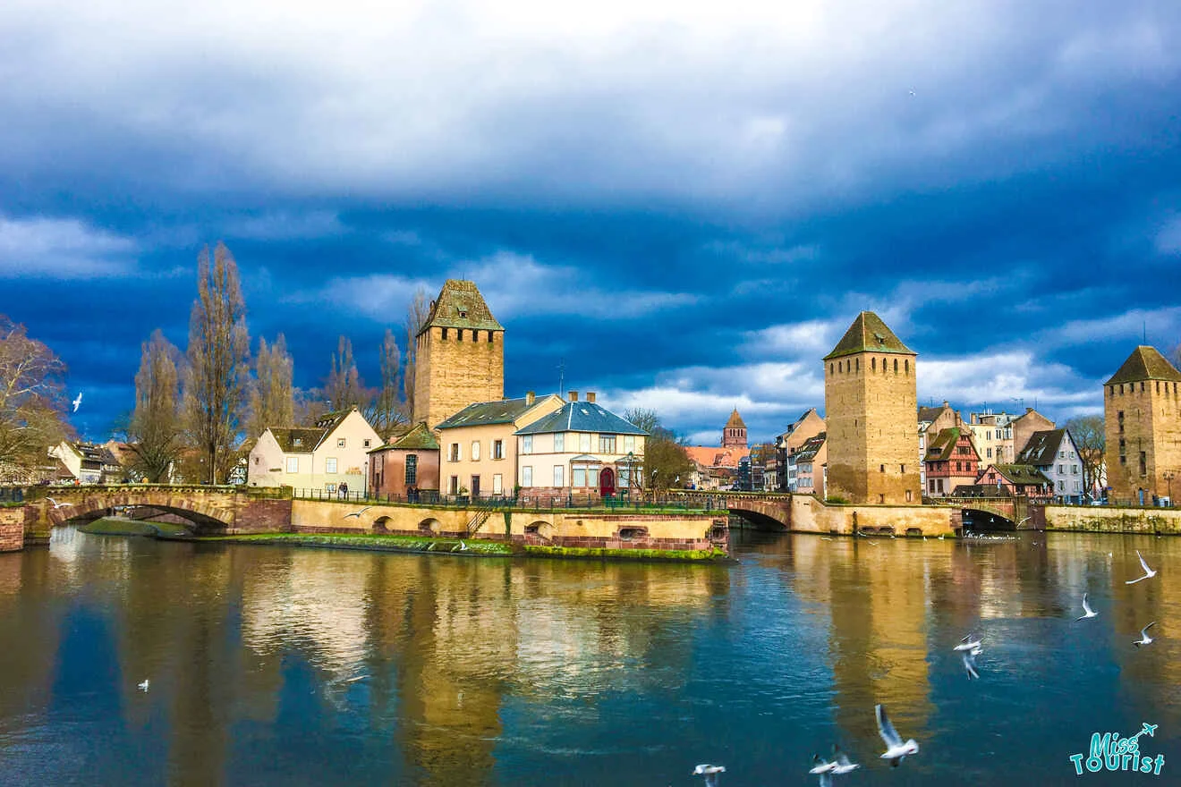 Historical towers and buildings reflect on a river under a dramatic cloudy sky, with seagulls flying nearby.