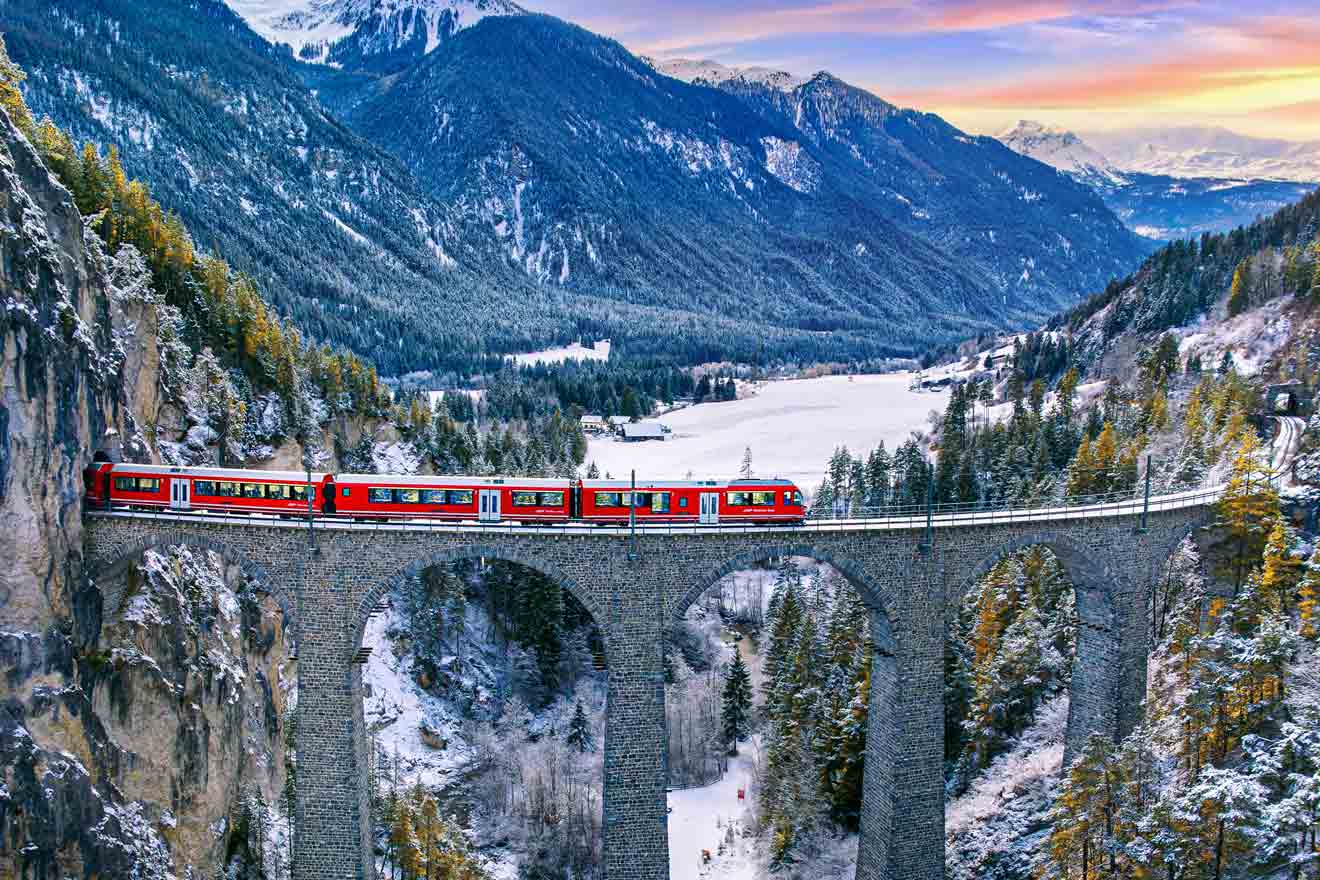 A red train crosses a stone viaduct amidst snow-covered mountains and forests under a colorful sky.