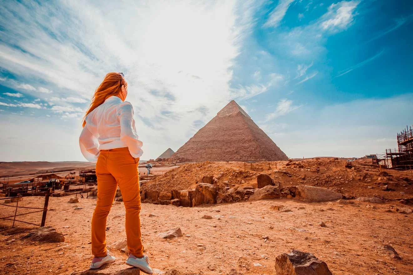 A person in a white shirt and orange pants stands facing the Great Pyramid of Giza under a blue sky with scattered clouds.