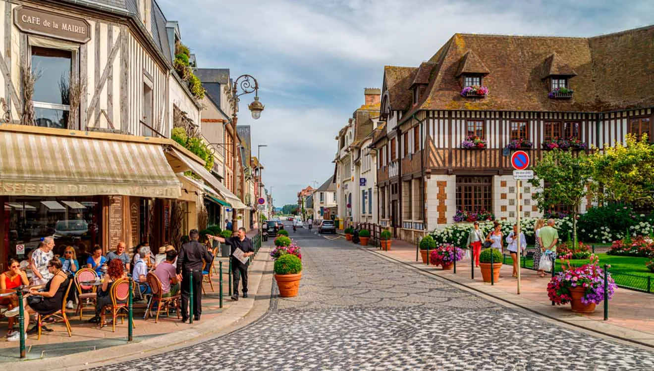 A quaint European street in Deauville with cobblestone road, outdoor cafe, and half-timbered buildings adorned with flowers. People are sitting at tables under a canopy.