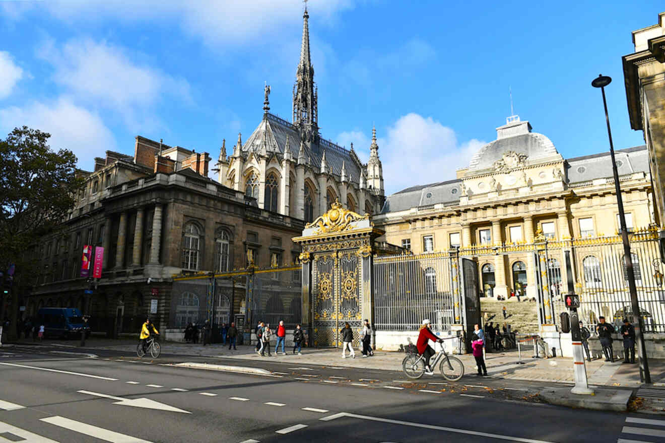 People walk and cycle on a street in front of a historic building with ornate gates, featuring Gothic architecture and a tall spire under a blue sky.