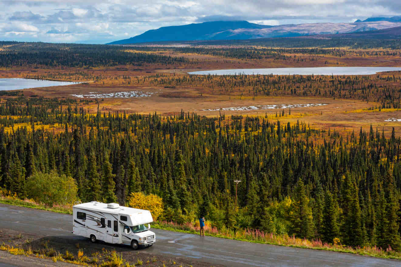 A white RV is parked on a roadside overlooking a vast landscape of trees and a distant body of water under a cloudy sky.