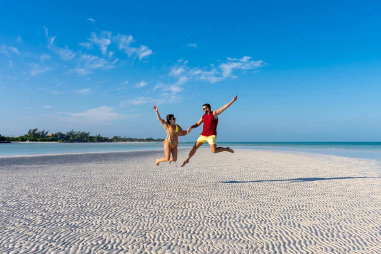 A man and a woman in summer clothing hold hands and jump on a sandy beach against a clear blue sky.