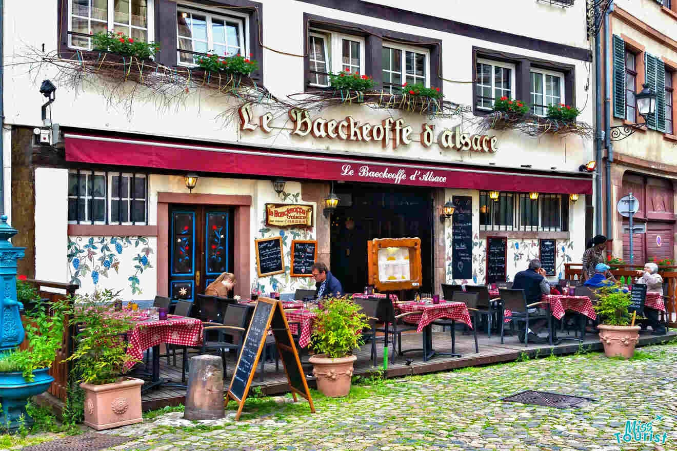 A quaint restaurant with outdoor seating, red checkered tablecloths, and patrons dining. The sign reads "Le Baeckeoffe d'Alsace." The building facade is decorated with plants and mosaics.