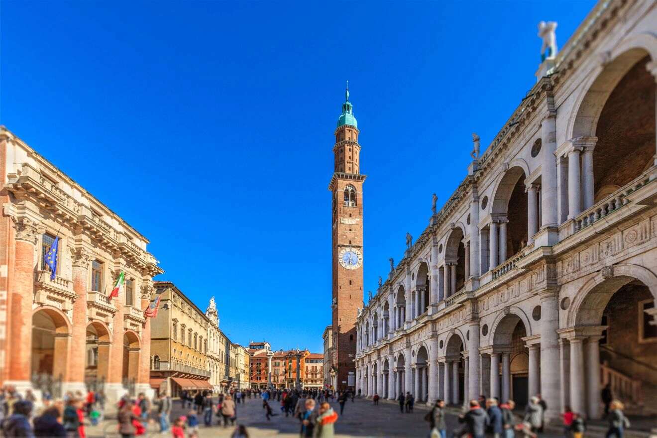 A tall clock tower stands in a historic square surrounded by Renaissance-style buildings, with people walking on a sunny day.