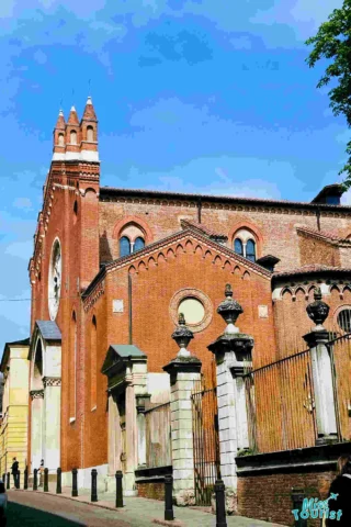 A historic red brick church with pointed arches and a tall bell tower, bordered by a stone fence, under a clear blue sky.