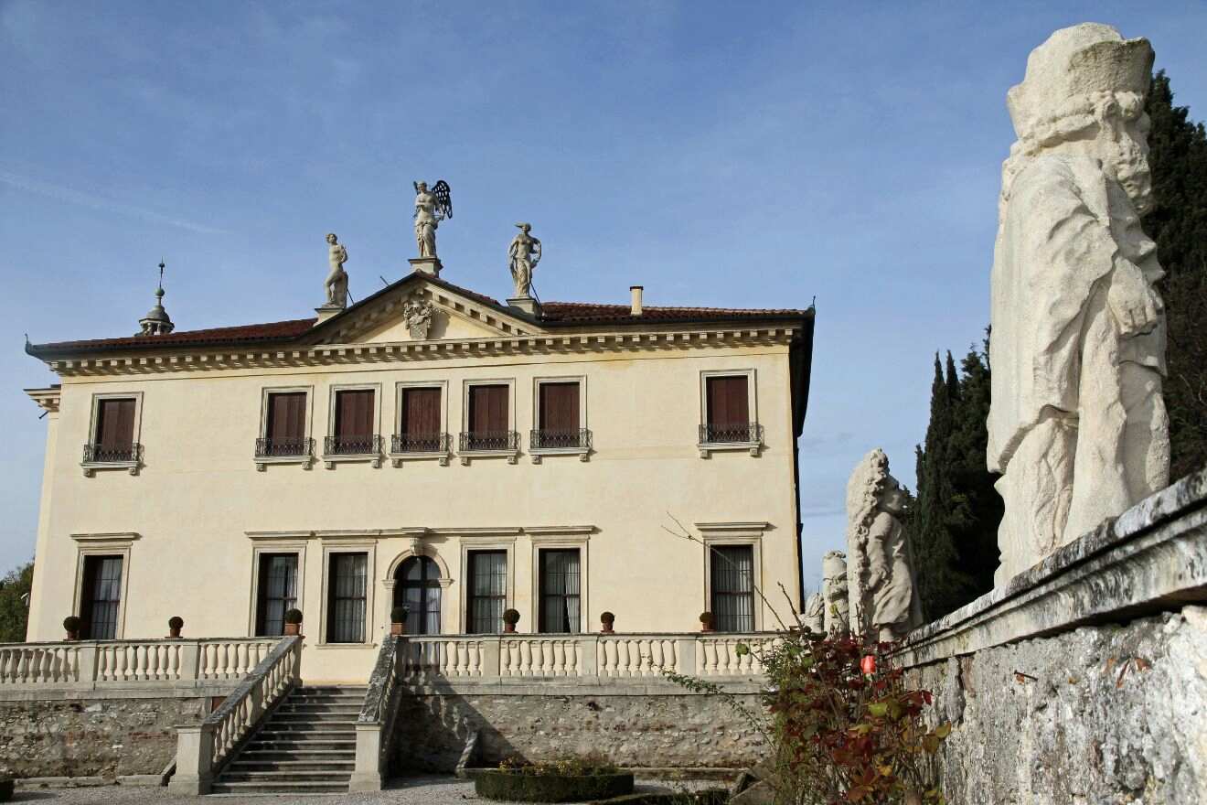 A historic villa with a symmetrical facade, arched entrance, and statues on the roof and along the entry stairway, set against a clear sky.