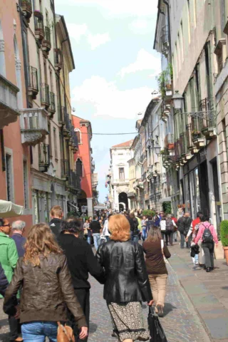 A busy pedestrian street lined with old buildings, with people walking in both directions under a cloudy sky.
