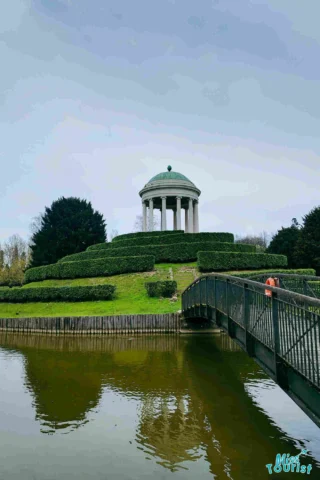 A domed stone pavilion on a hedge-covered mound, with a footbridge crossing a pond in the foreground under an overcast sky.