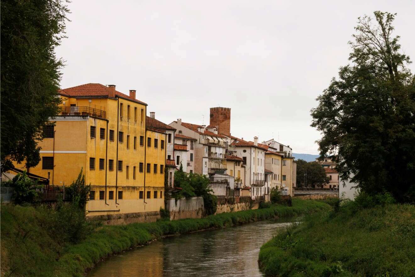 Row of colorful buildings, including a prominent yellow one, lines a narrow river with greenery on both sides under an overcast sky.