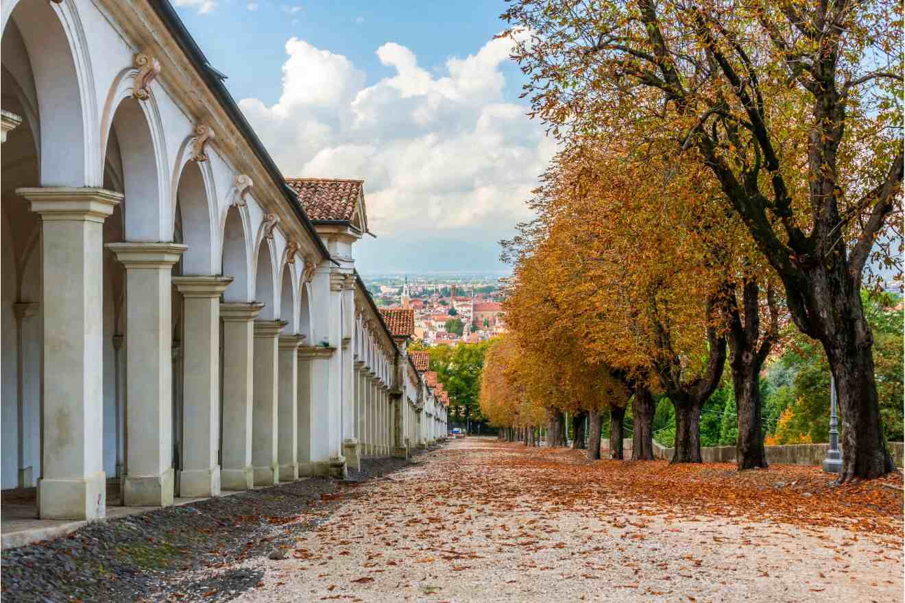 A tree-lined walkway with autumn leaves runs parallel to a colonnaded building, overlooking a distant cityscape under a partly cloudy sky.