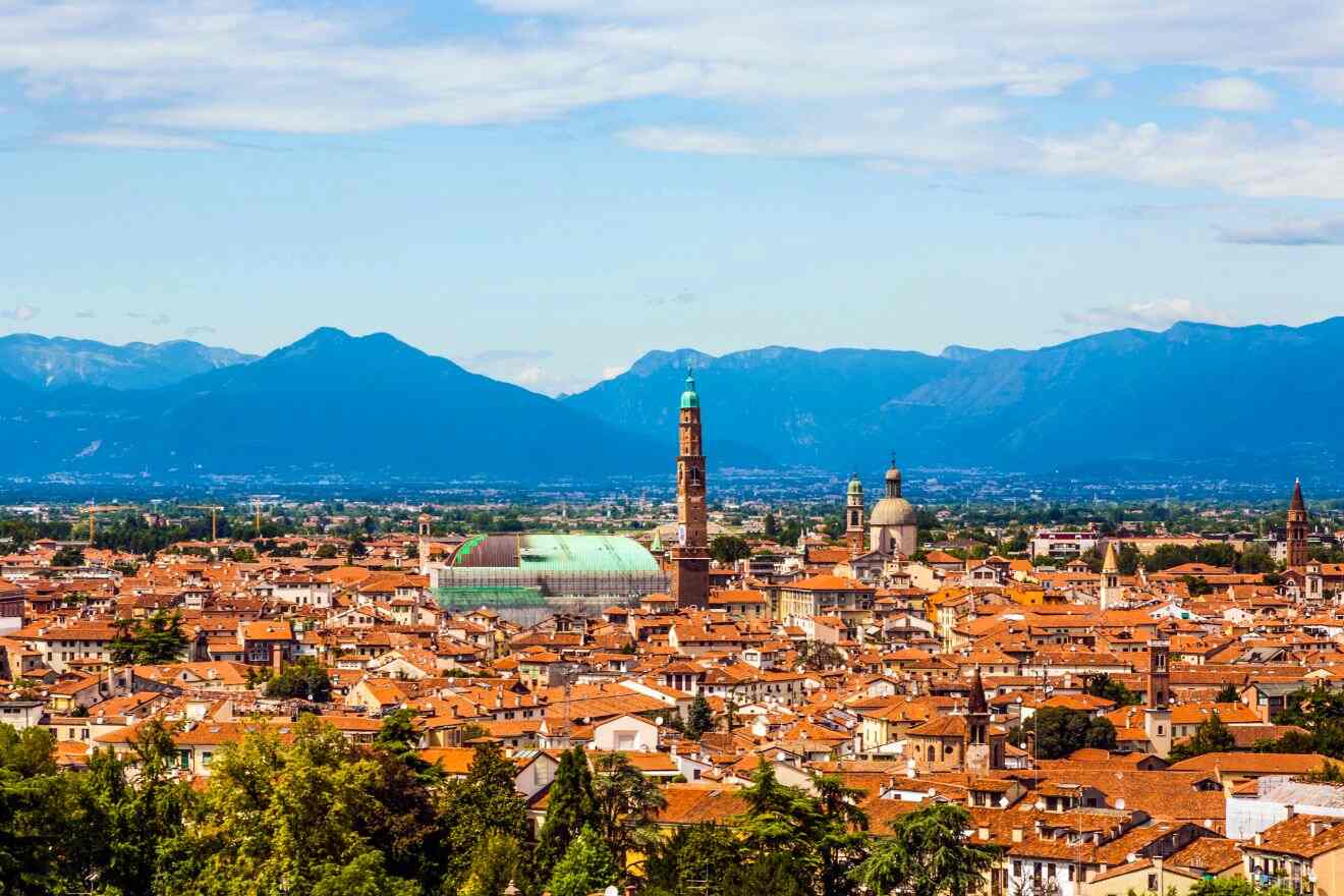 A panoramic view of a city with red-tiled roofs, a tall clock tower, a domed building, and mountains in the background under a partly cloudy sky.