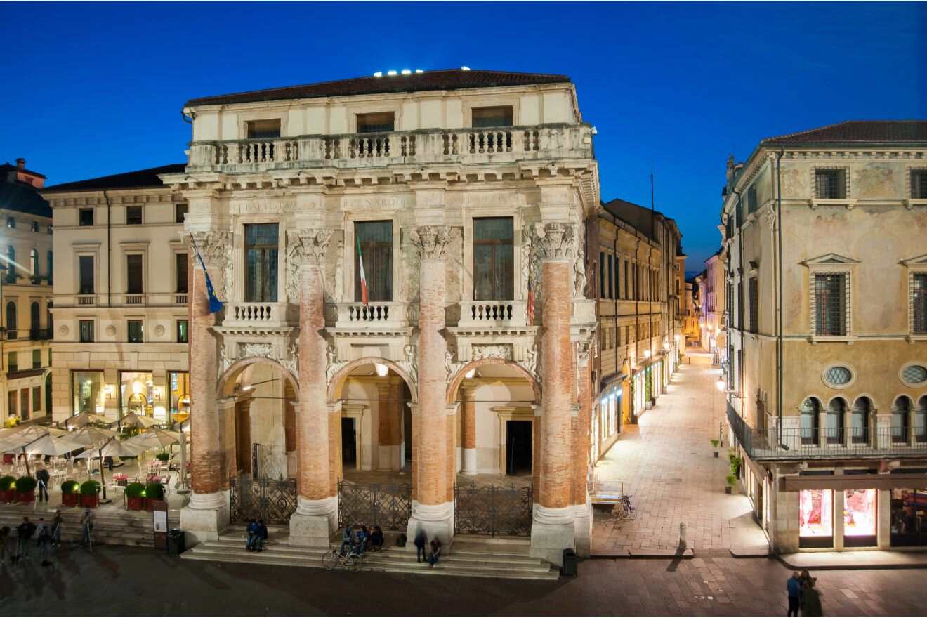Historic stone building with columns and arched entrances, illuminated at dusk, stands at the intersection of two pedestrian streets in a European city.