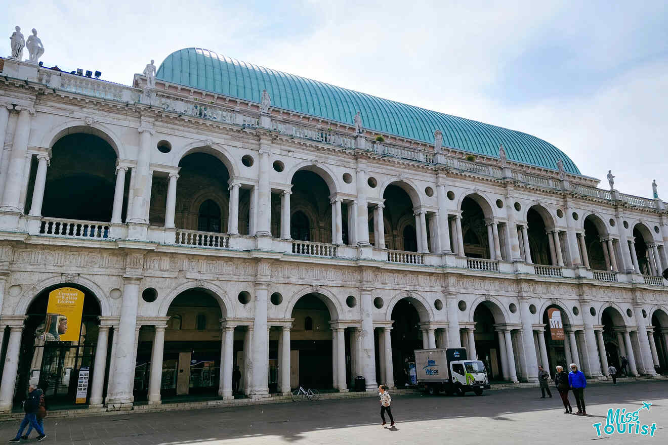 A historic white stone building with arched windows and columns, featuring a green dome roof and people walking in the plaza below.