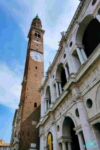 A tall brick clock tower stands next to a white stone building with arches and columns under a partly cloudy sky.