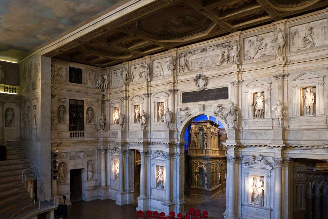 Interior of a historic theater with ornate stone arches, classical statues, reliefs, and a blue-lit stage seen through a central archway. Red chairs are arranged in front.