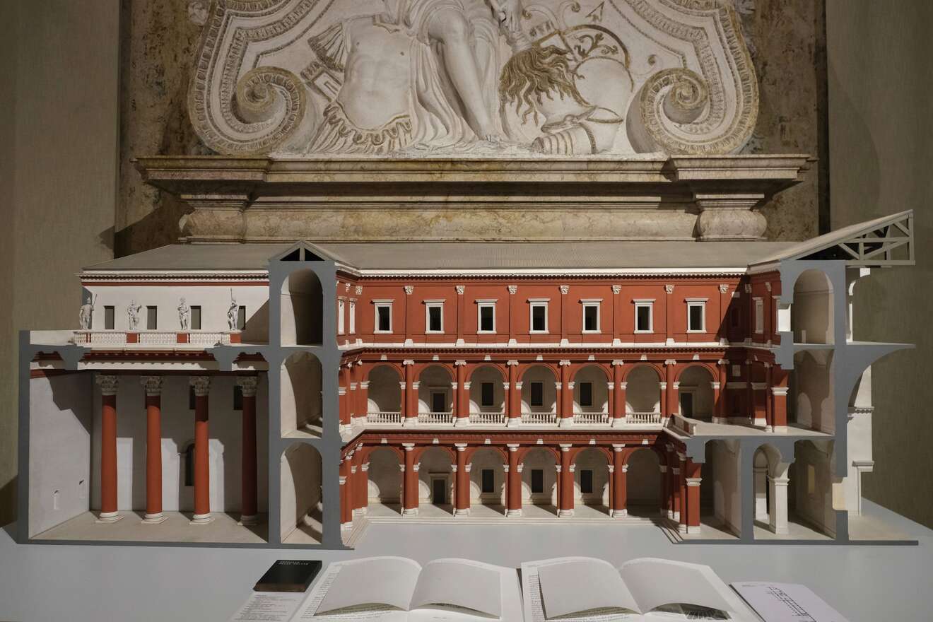 A detailed architectural model of a classical building with columns and arches is displayed in front of a carved stone relief; open books lie on the table below.