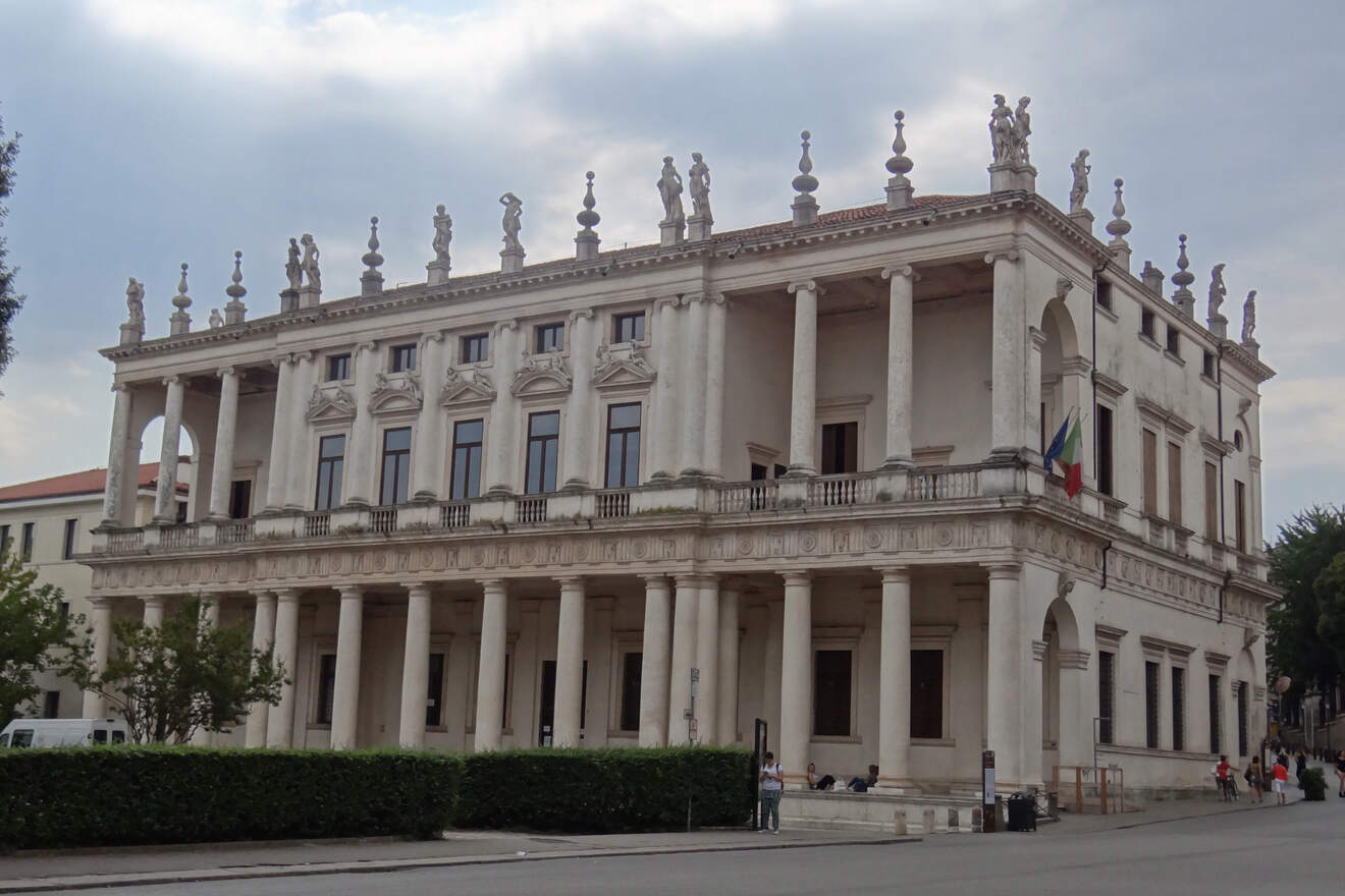 A large, ornate neoclassical building with columns, statues on the roof, and Italian flags at the entrance, set against a cloudy sky.