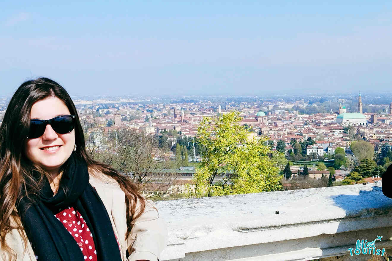 The writer of the post wearing sunglasses and a scarf poses in front of a scenic city view with historic buildings and a clear sky in the background.