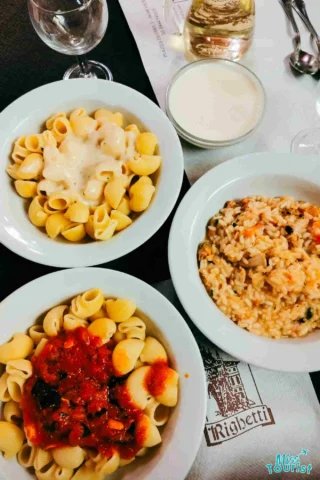 Three bowls of Italian pasta and risotto dishes on a table, served with a glass of white wine and an empty water glass.
