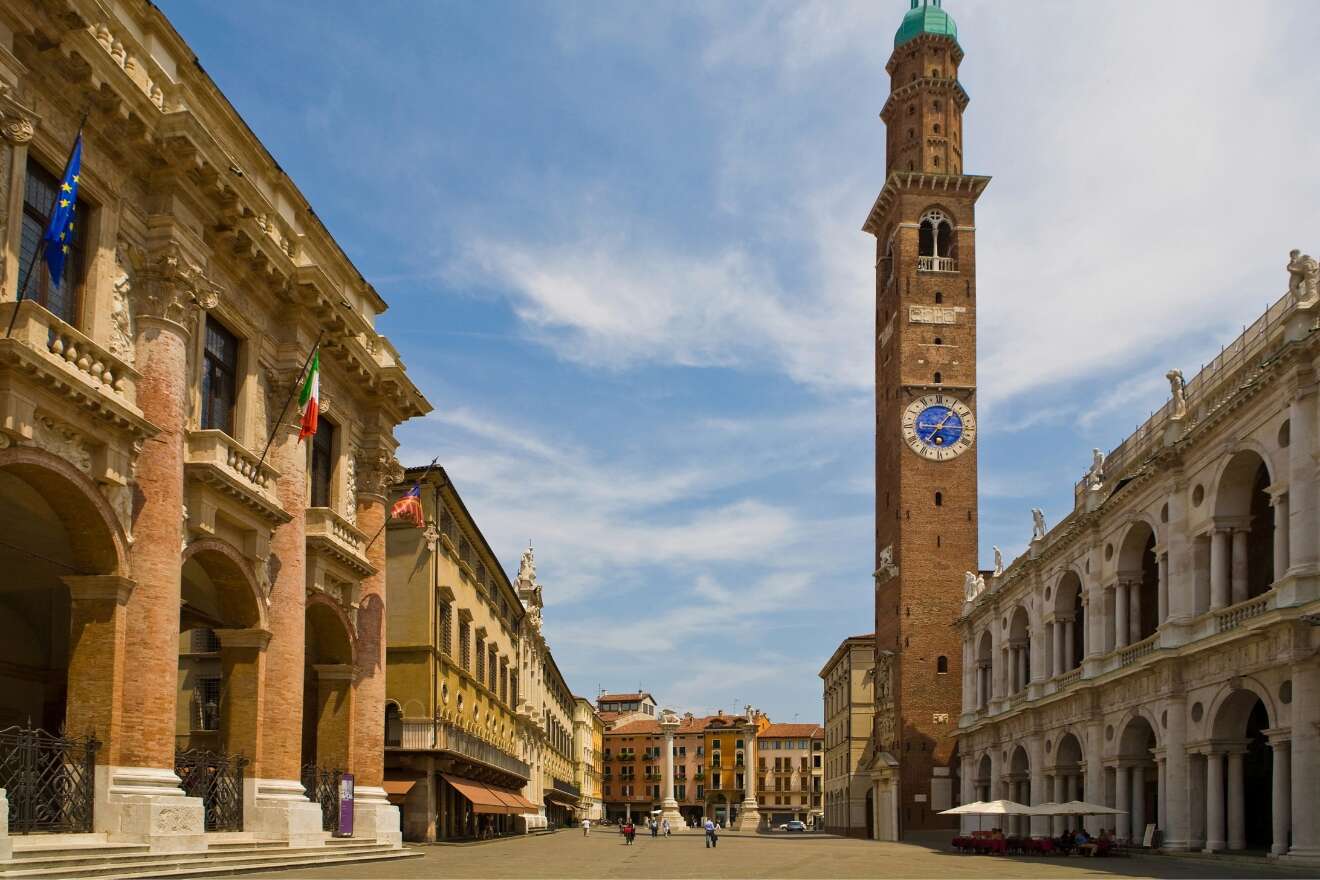 A tall brick clock tower stands in an open square surrounded by historic buildings with arches and columns under a blue sky.