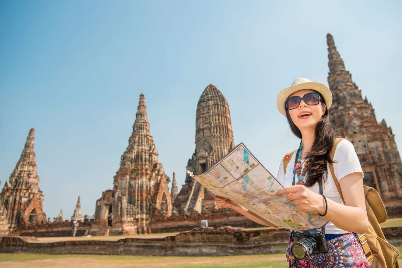 A woman wearing a hat and sunglasses holds a map in front of ancient temple ruins, with tall stone spires in the background under a clear blue sky.