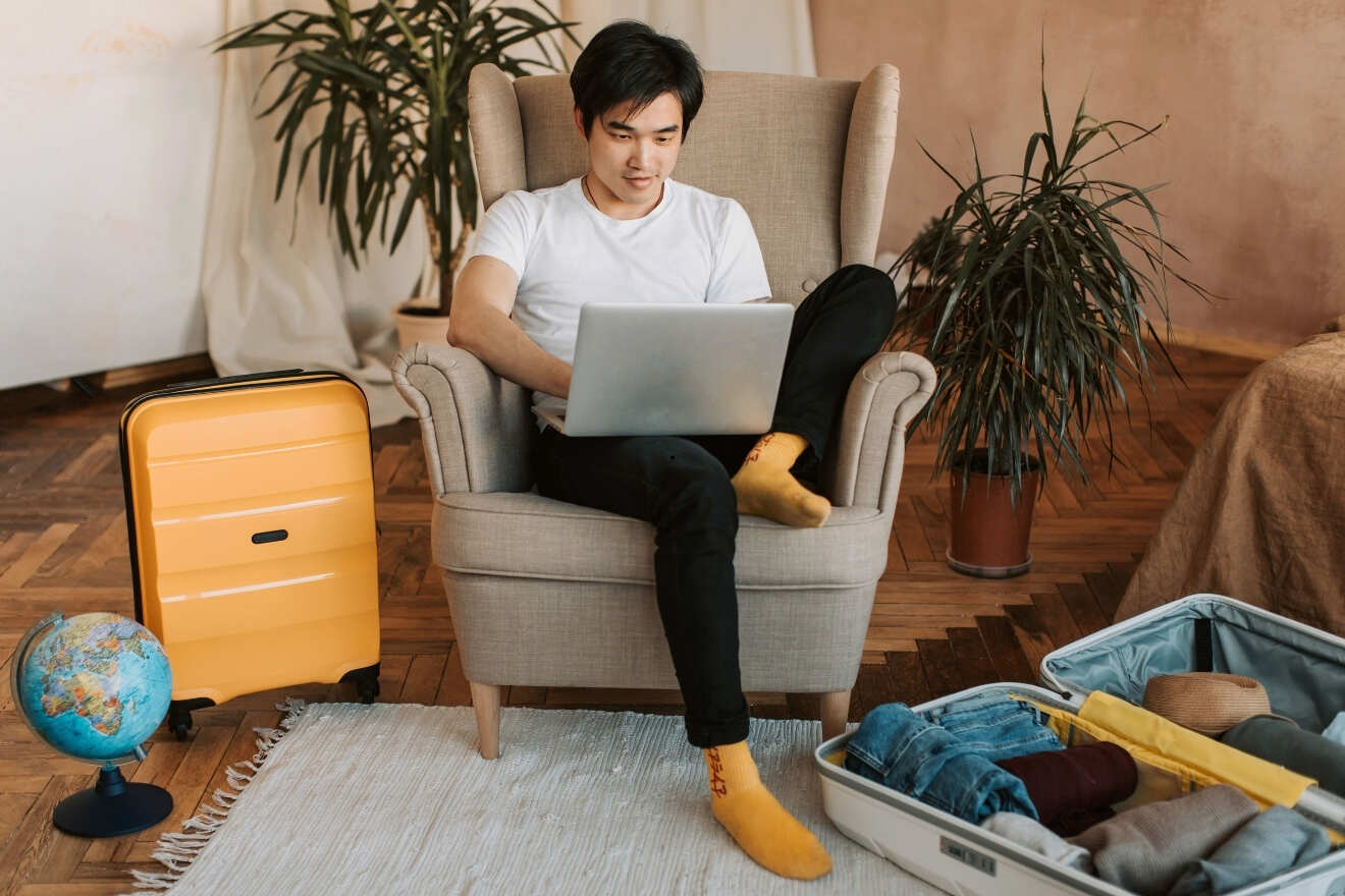 Person sitting on an armchair using a laptop, with a yellow suitcase, open luggage, and a globe nearby in a cozy room with plants.