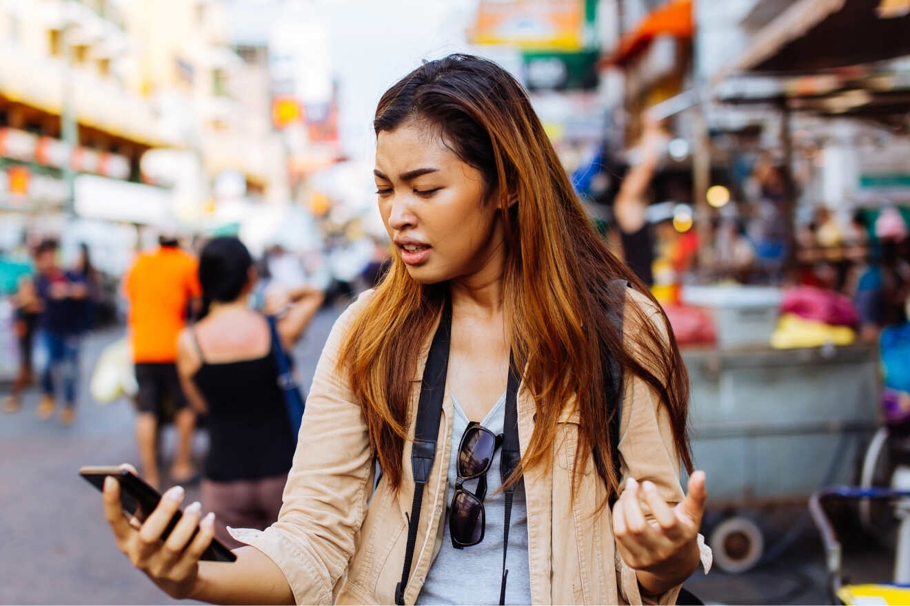 A woman standing on a busy street looks at her phone with a confused expression, holding it in one hand and gesturing with the other.