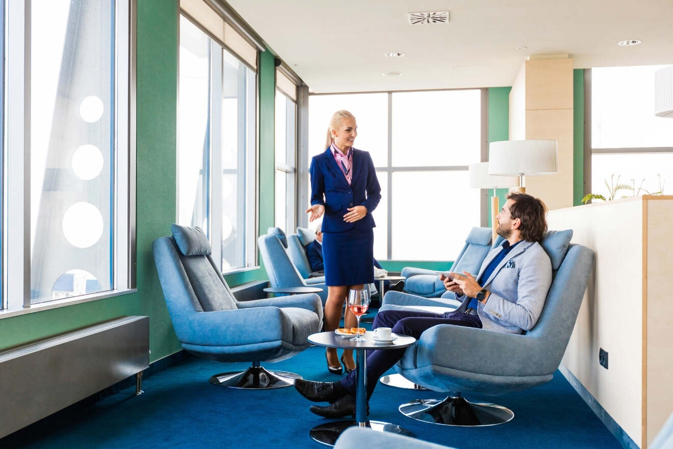 A woman in a blue uniform stands beside a seated man in a lounge area with large windows, blue chairs, and drinks on small tables.
